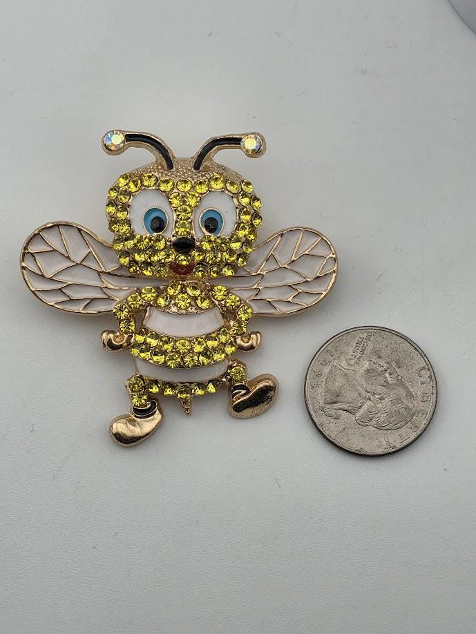 A yellow and gold rhinestone-encrusted brooch in the shape of a bee is placed next to a US quarter on a white surface.