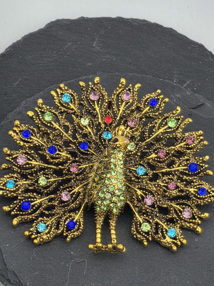 A decorative gold peacock brooch with colorful gemstones, including blue, green, pink, and purple stones, set against a dark background.