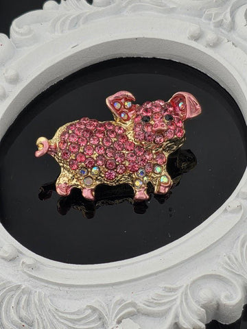 A pink and gold rhinestone-encrusted brooch in the shape of a pig is displayed against a black background.