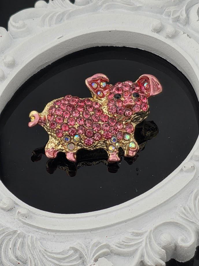 A pink and gold rhinestone-encrusted brooch in the shape of a pig is displayed against a black background.