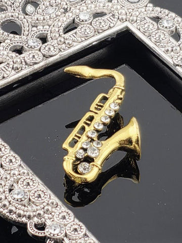 A gold saxophone pendant with rhinestones is displayed on a black surface, with a silver ornate frame in the background.