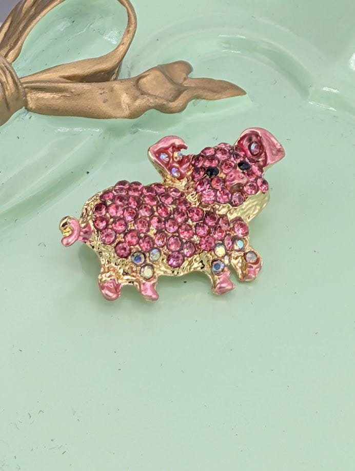 A pink and gold rhinestone-encrusted brooch in the shape of a pig is displayed against a black background.