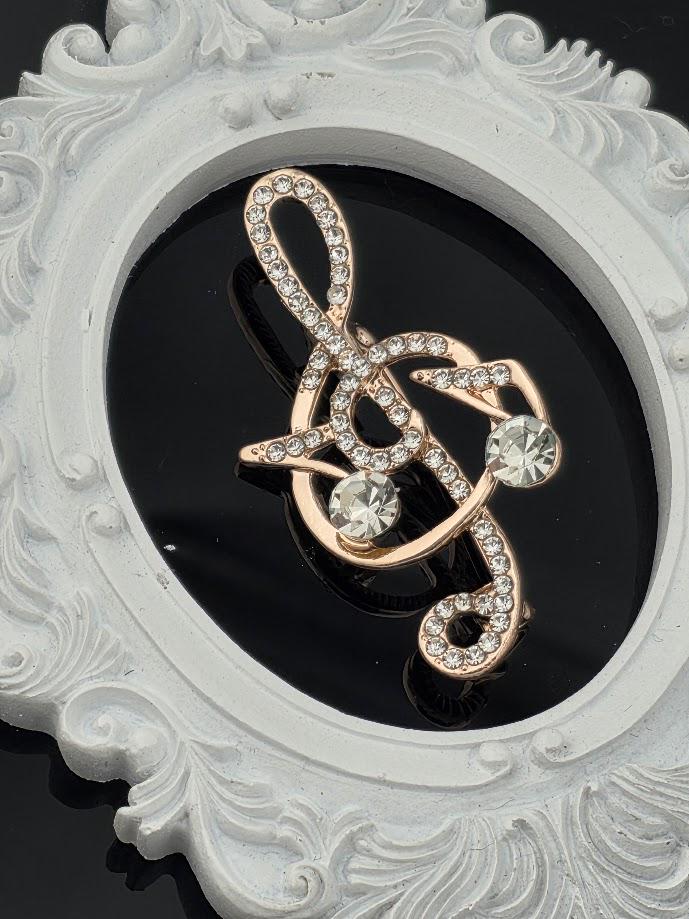 A decorative brooch in the shape of a treble clef, adorned with sparkling rhinestones, is displayed against a black background within an ornate white frame.