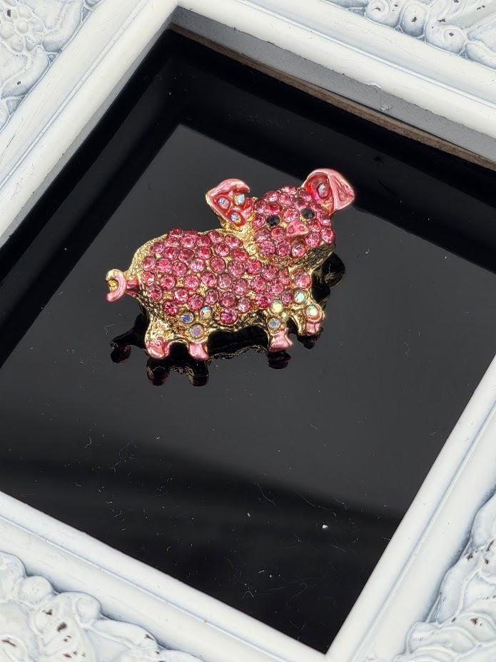 A pink and gold rhinestone-encrusted brooch in the shape of a pig is displayed on a black surface, framed by a white border.