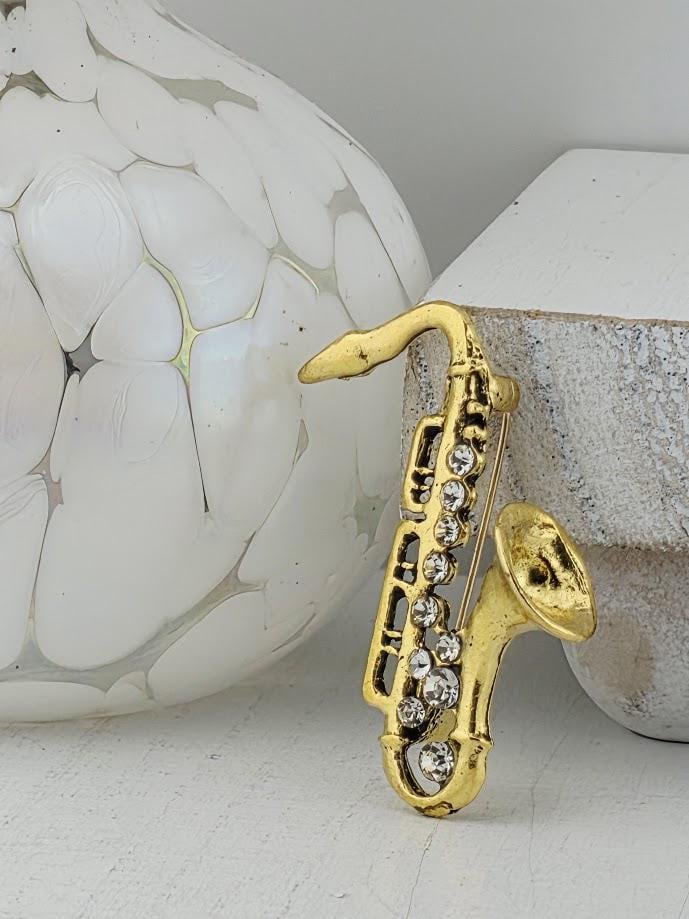 A gold-colored saxophone brooch with rhinestones is displayed against a white background, accompanied by a white vase and a white ceramic object.