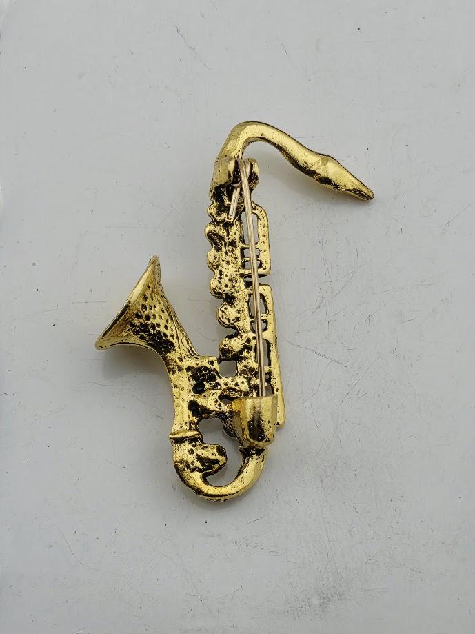 A gold-colored saxophone brooch is shown against a white background.