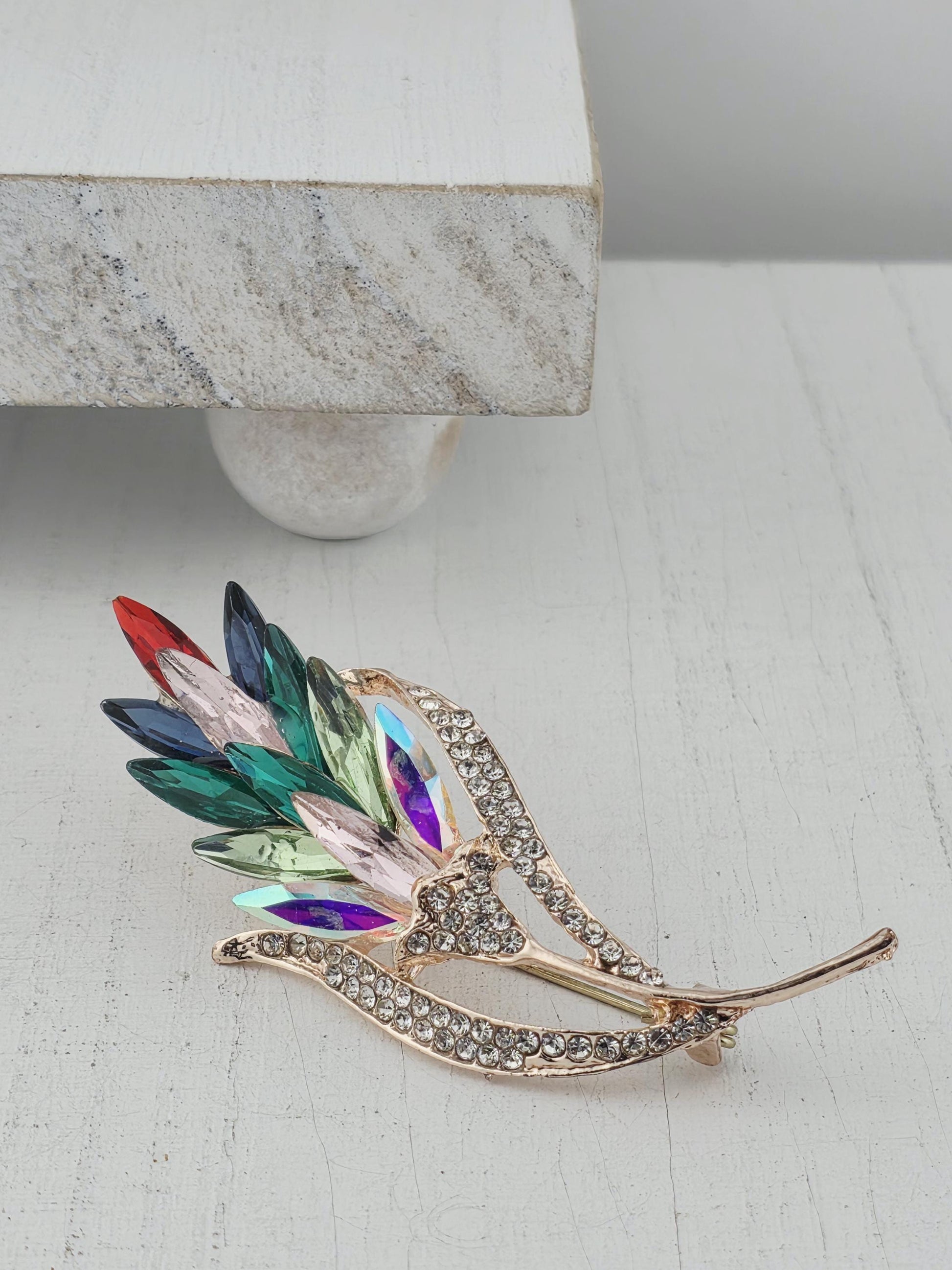 A decorative brooch with a leaf-like design and colorful gemstones is displayed against a white wall.