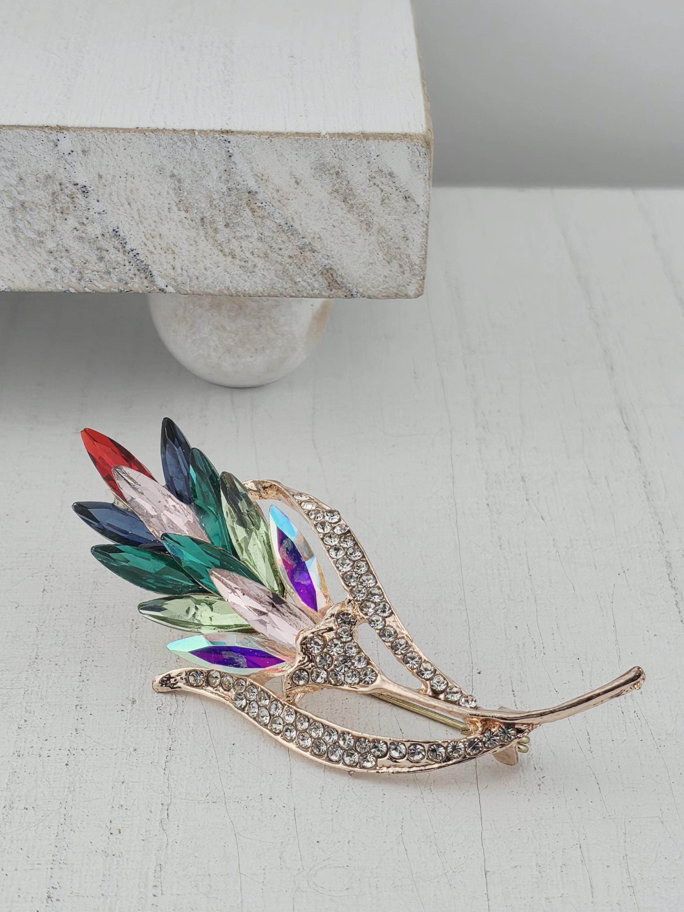 A decorative brooch with a leaf-like design and colorful gemstones is displayed against a white wall.