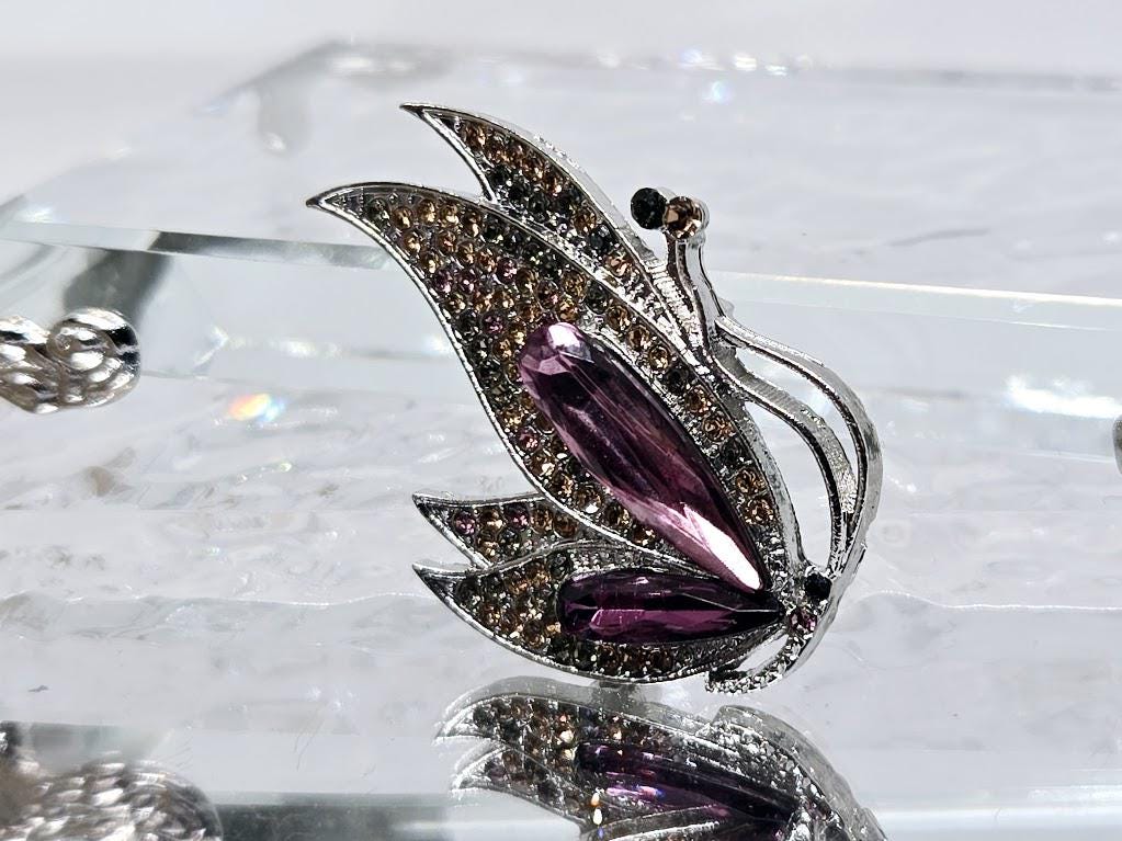 A decorative butterfly-shaped pendant with a purple gemstone on its wing, set against a reflective surface.
