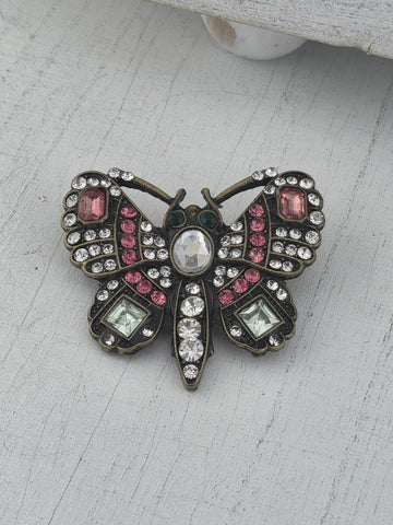 A decorative butterfly brooch with a combination of pink and green gemstones, along with a central stone, is displayed on a white surface.
