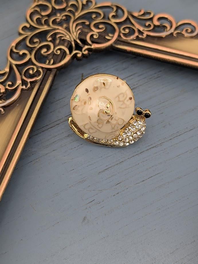 A decorative brooch in the shape of a snail, adorned with rhinestones, is displayed on a wooden surface.