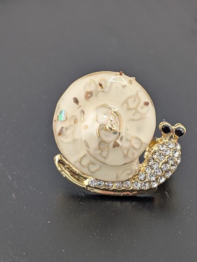 A decorative pin or brooch in the shape of a snail, with a white shell and a gold body adorned with rhinestones or crystals.