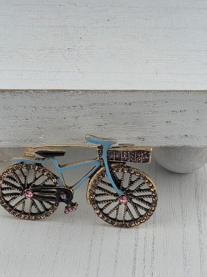 A decorative bicycle brooch with a blue frame and gold accents, featuring a pink gemstone on the front wheel.