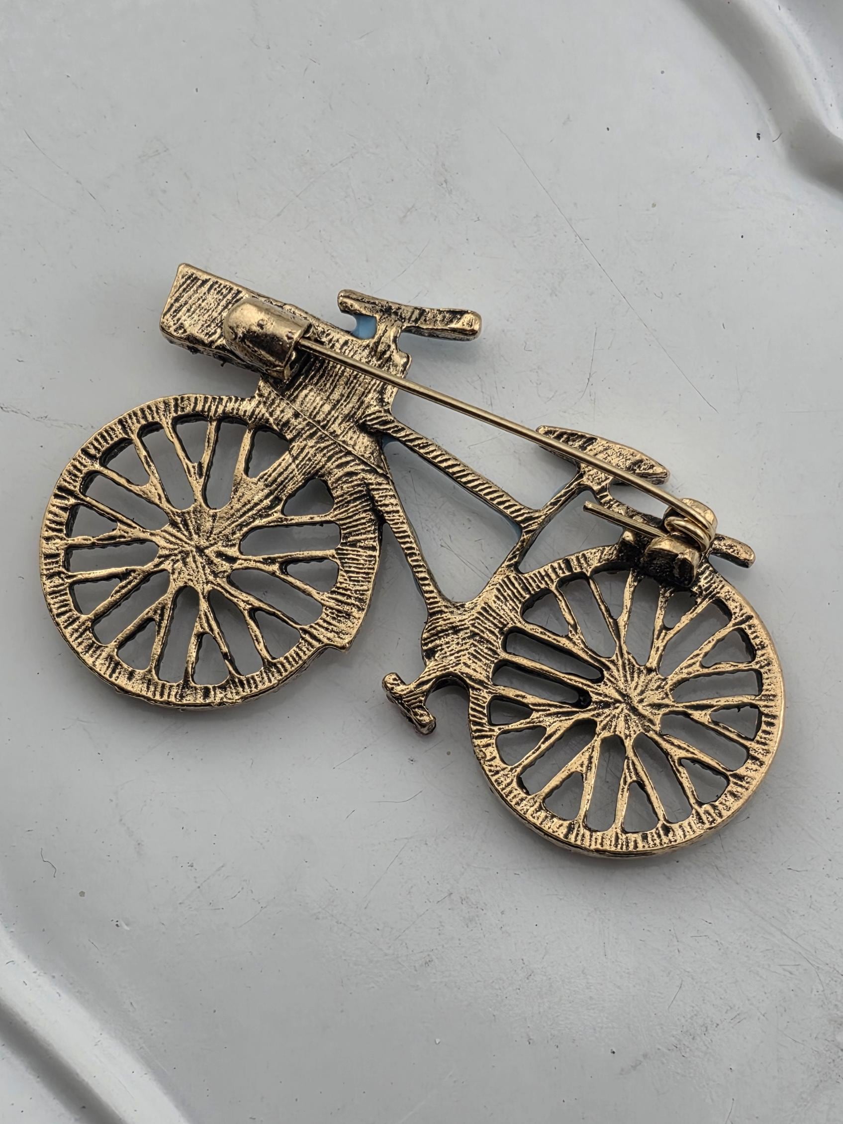 A golden, intricately designed bicycle brooch or pin is shown against a plain white background.