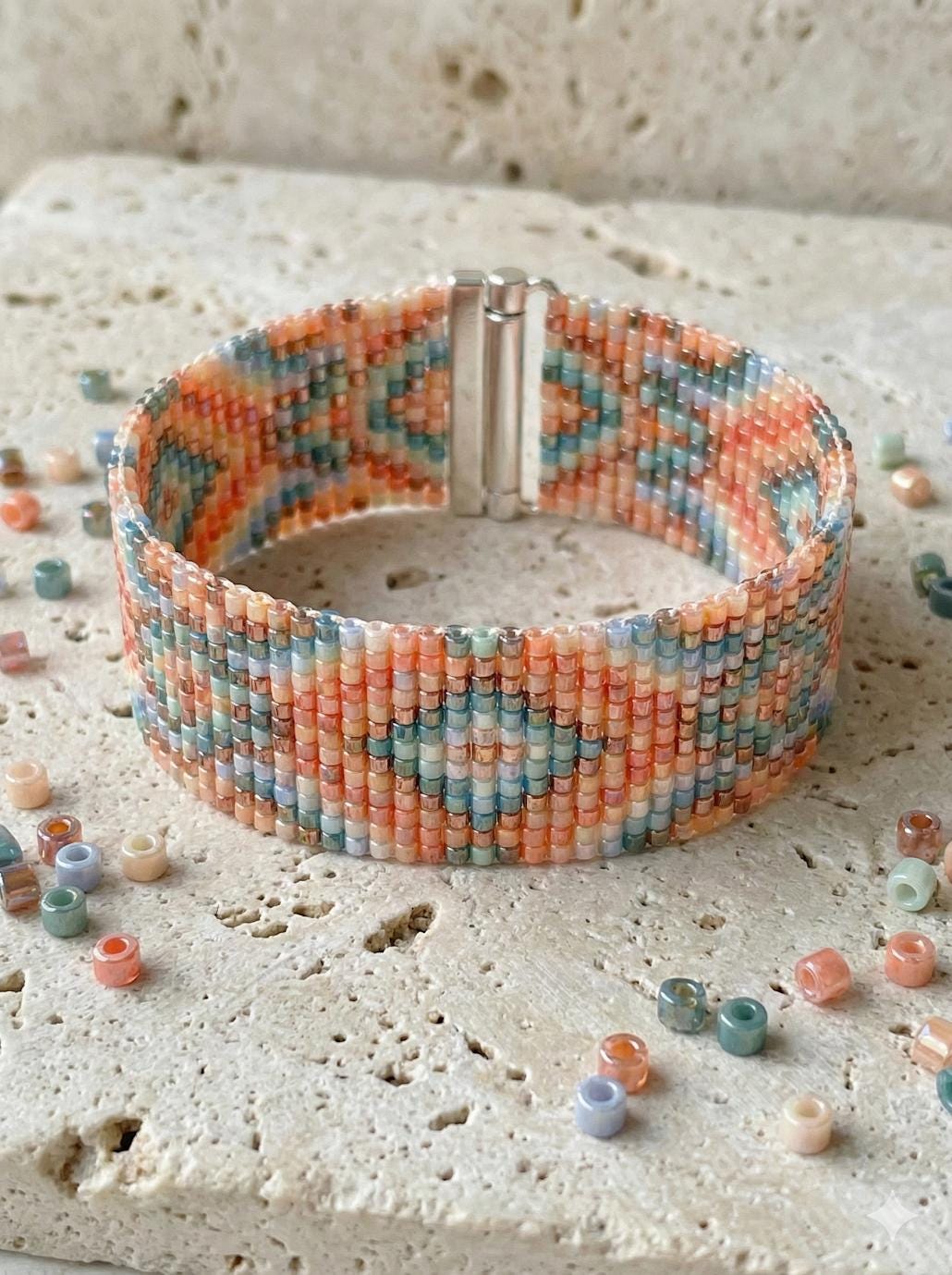 A beaded bracelet with a colorful, geometric pattern of small beads in various shades of orange, blue, and green. The bracelet is placed on a surface with scattered beads around it.