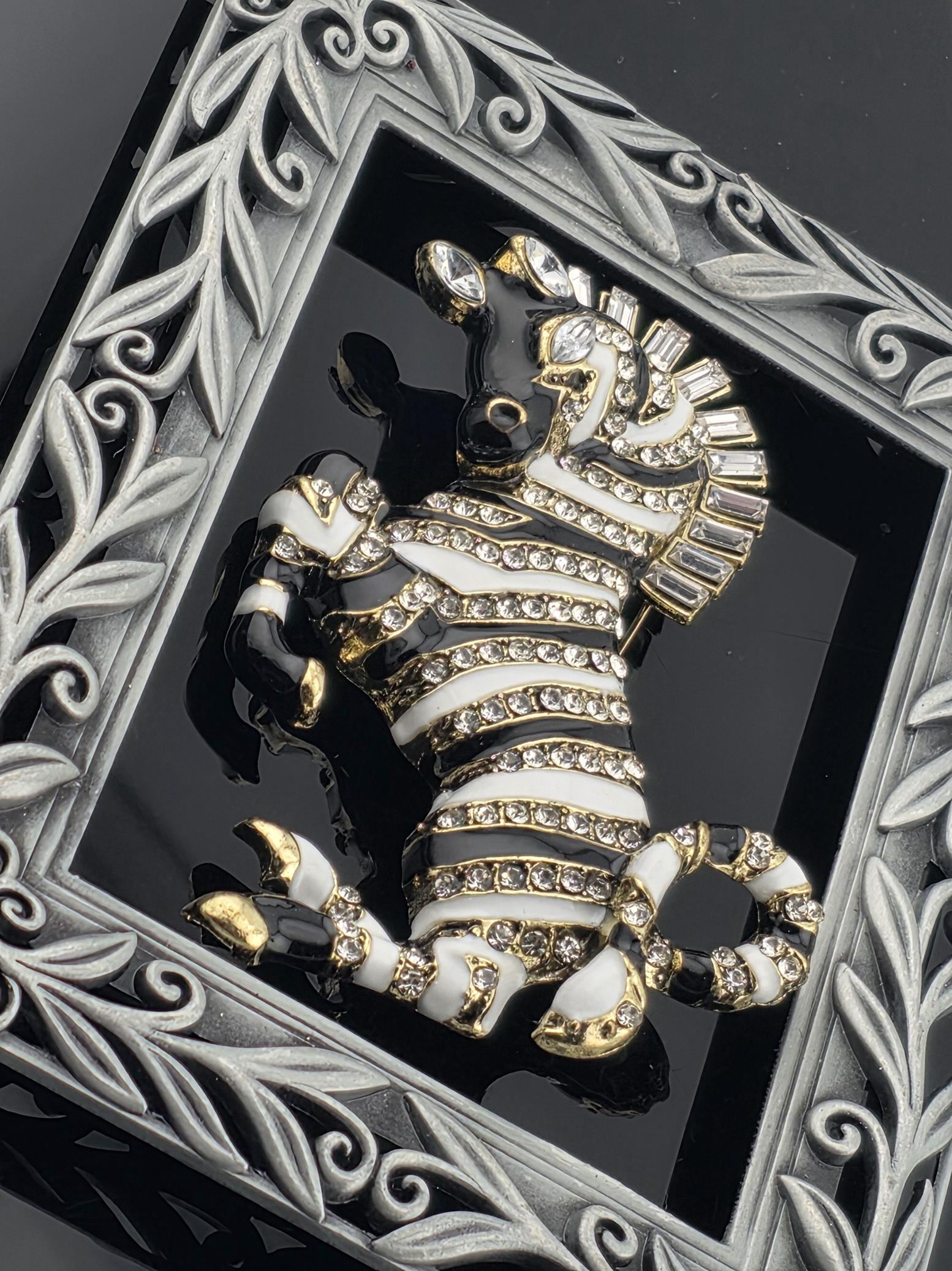 A decorative zebra brooch with a black and white striped pattern, adorned with rhinestones and placed within an ornate silver frame.