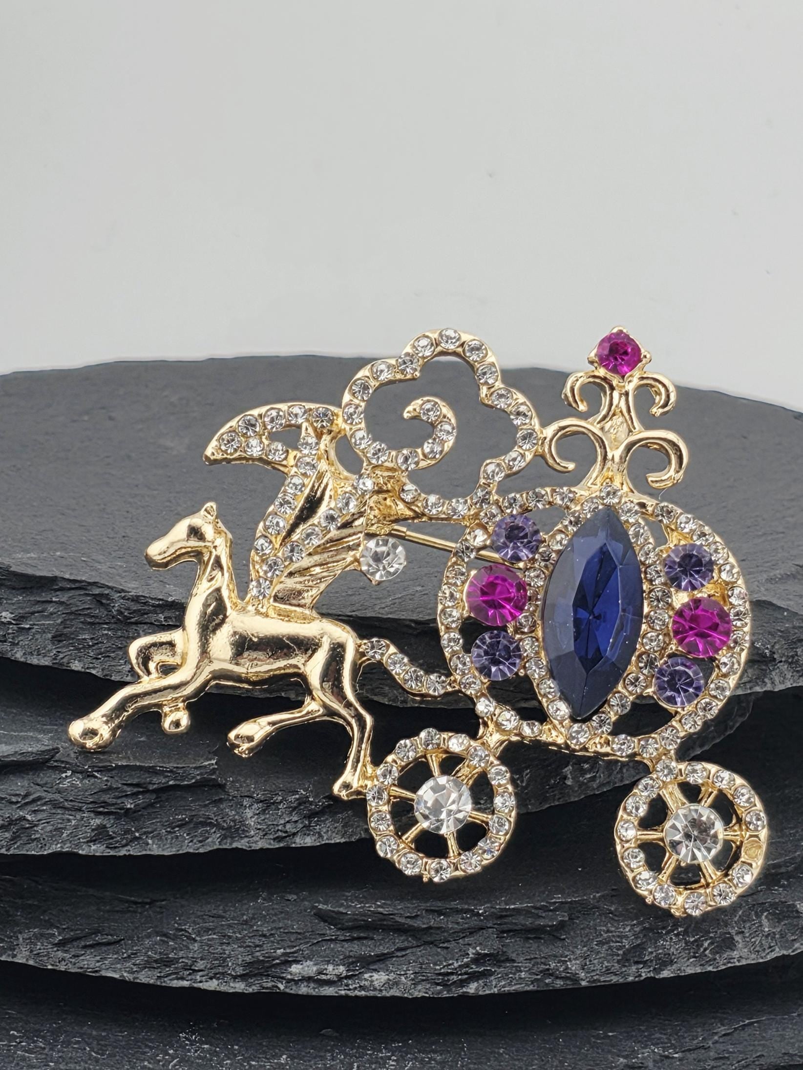 A decorative brooch with a gold-colored horse and various gemstones, including a blue gemstone, is displayed against a black background.