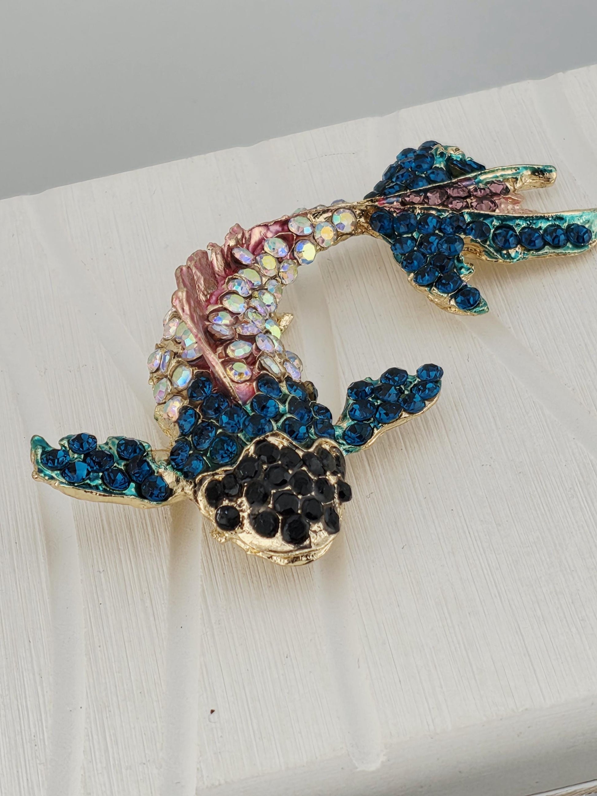 A decorative brooch in the shape of a fish, adorned with blue and pink gemstones, is displayed on a white surface.