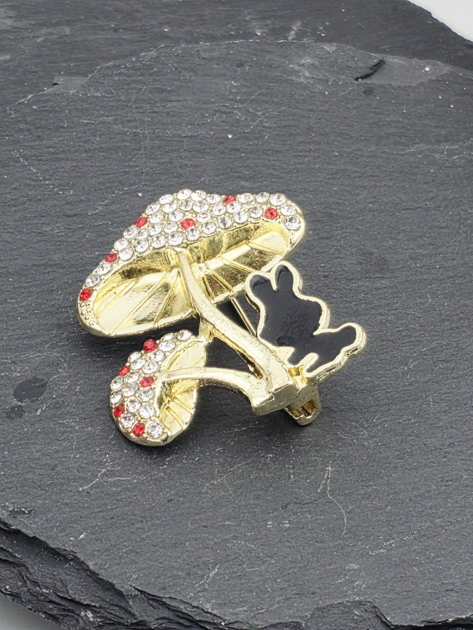 A decorative brooch with a stylized design, featuring a combination of gold and red gemstones, is displayed on a dark gray surface.