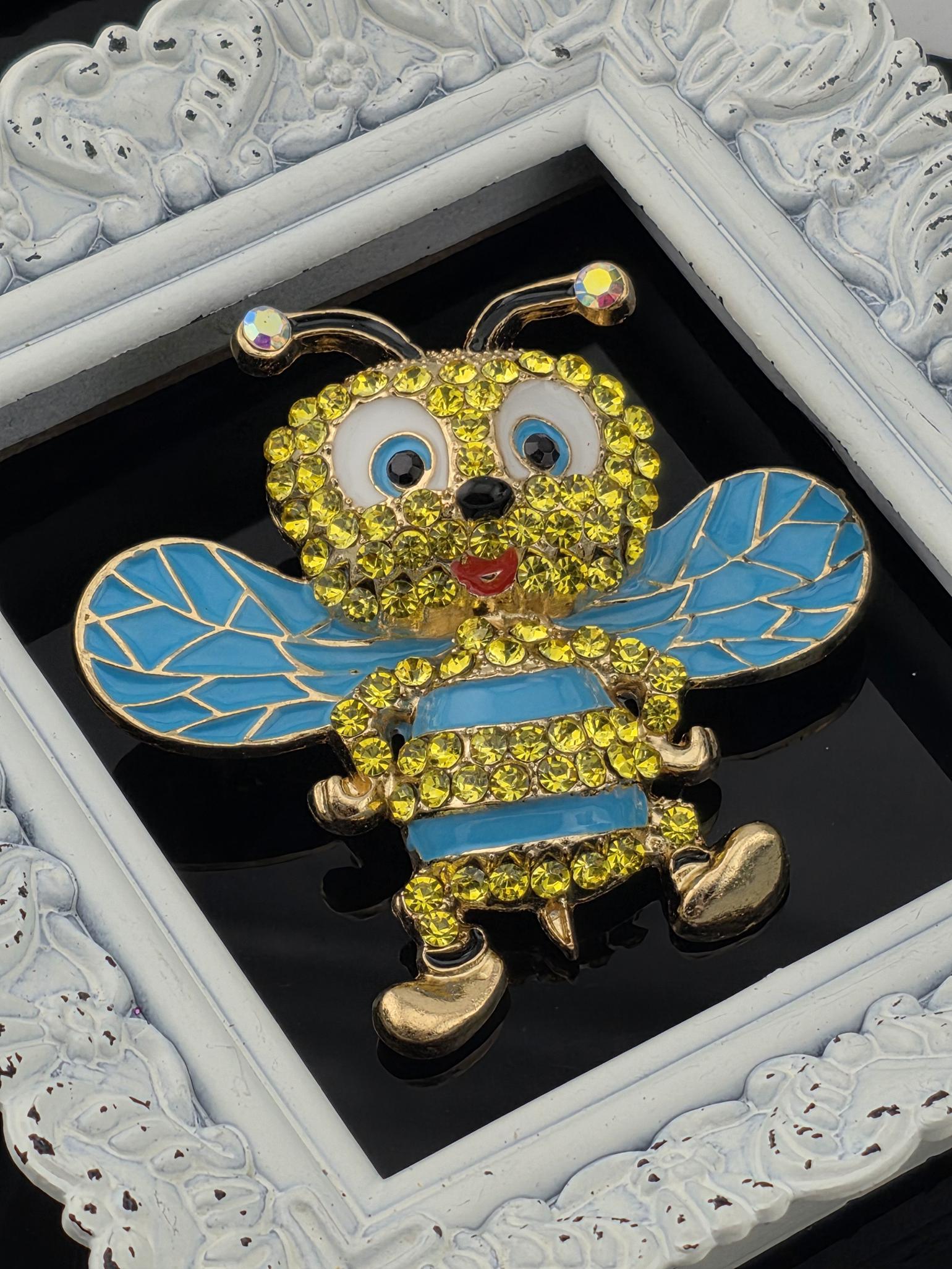 A decorative piece featuring a bee-like figure with a blue body, yellow accents, and a red tongue, set against a black background and framed by a white ornate frame.