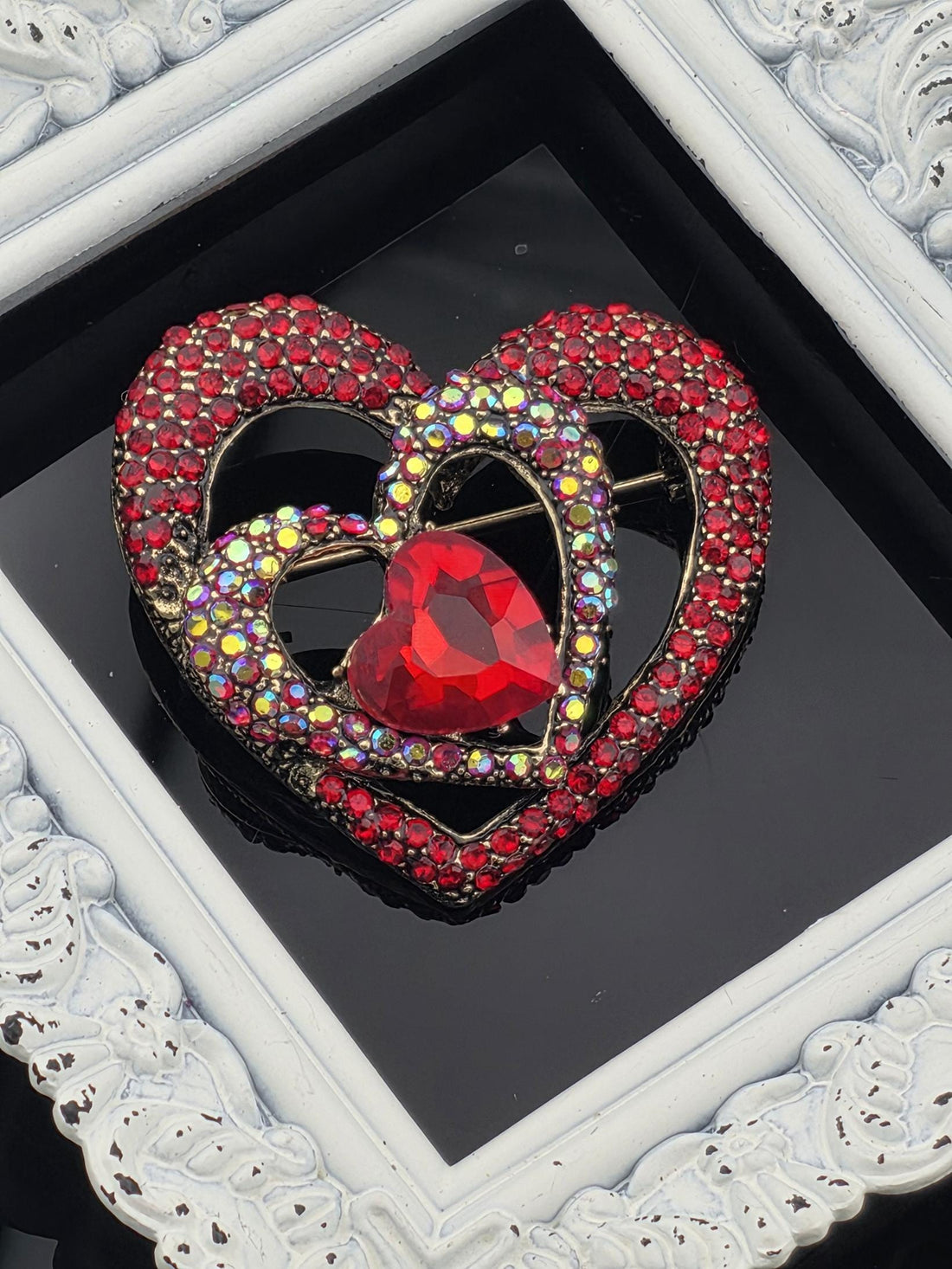 A red heart-shaped brooch adorned with sparkling rhinestones and a large red gemstone is displayed against a black background.