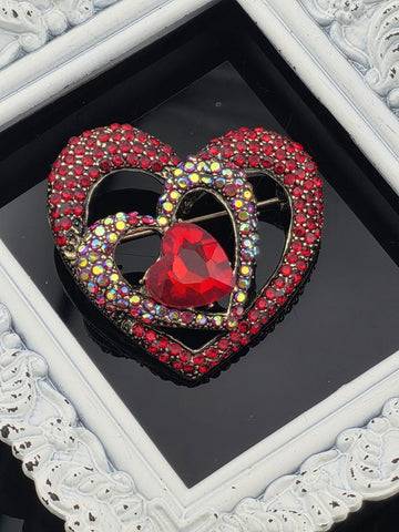 A red heart-shaped brooch adorned with sparkling rhinestones and a large red gemstone is displayed against a black background.