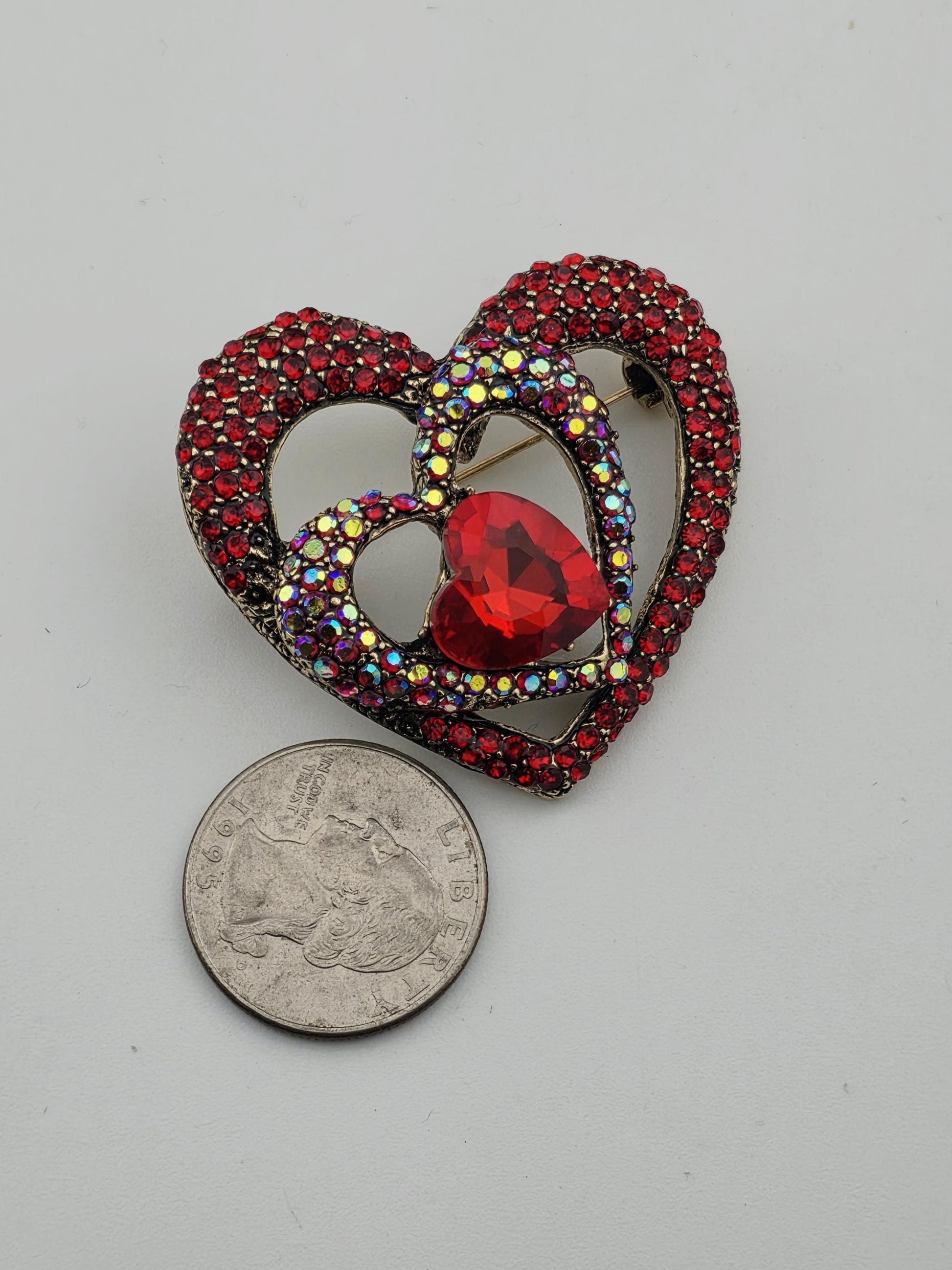 A heart-shaped brooch adorned with red gemstones and a large red gemstone is placed next to a quarter on a white surface.