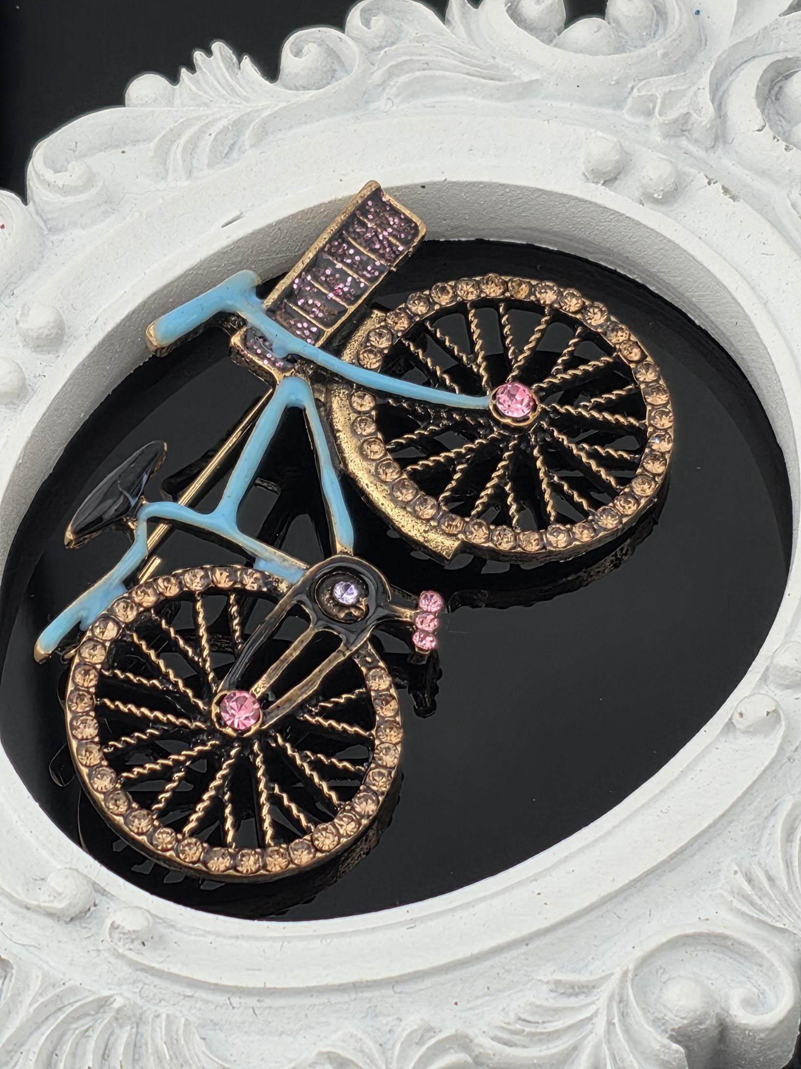A decorative bicycle with a blue frame and gold accents, including rhinestones and a pink gemstone, is displayed in a white ornate frame.