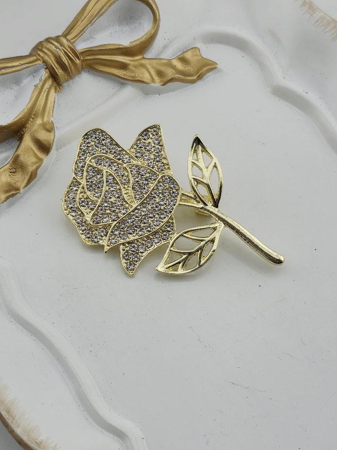 A gold-colored brooch with a diamond-encrusted rose design is displayed on a white surface, accompanied by a gold ribbon bow.