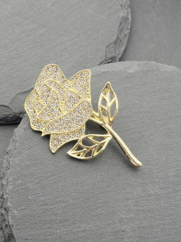 A gold-colored brooch with a diamond-encrusted rose design is displayed on a gray surface.