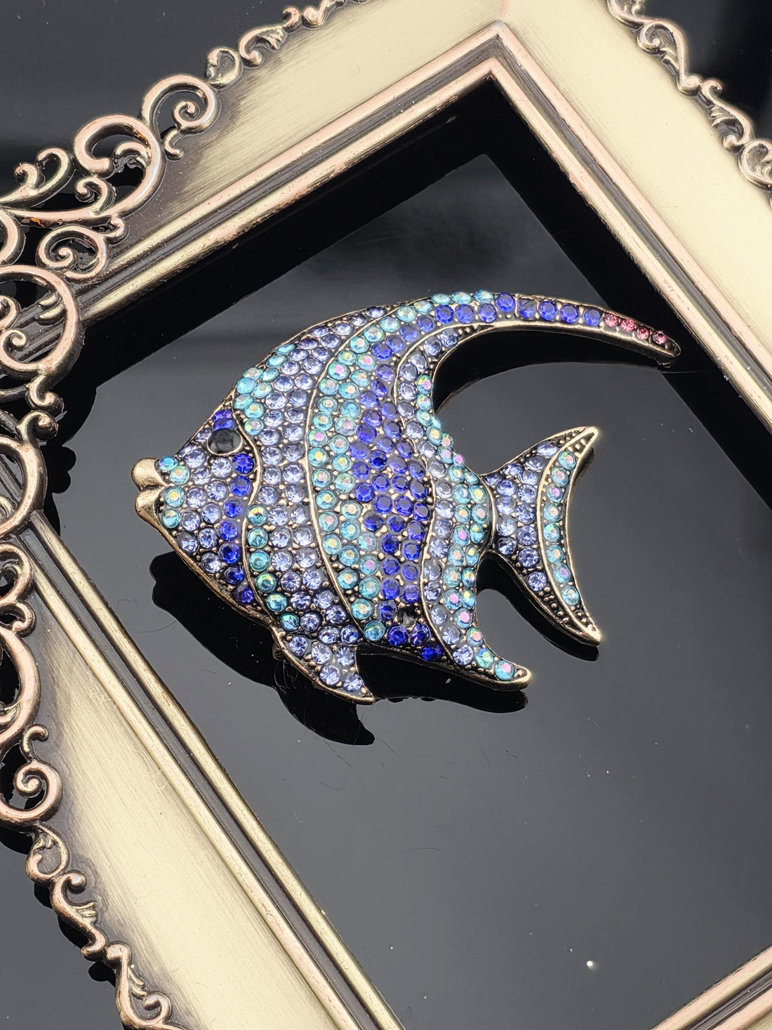 A decorative fish brooch with blue and purple gemstones is displayed in a gold frame.