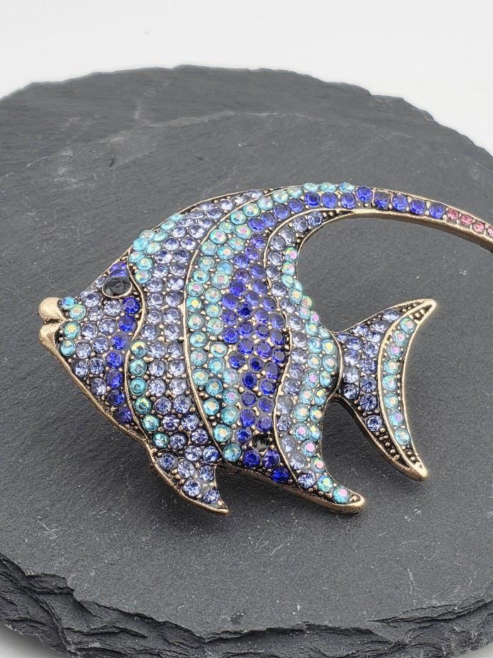 A decorative fish brooch with blue and purple gemstones is displayed in a gold frame.