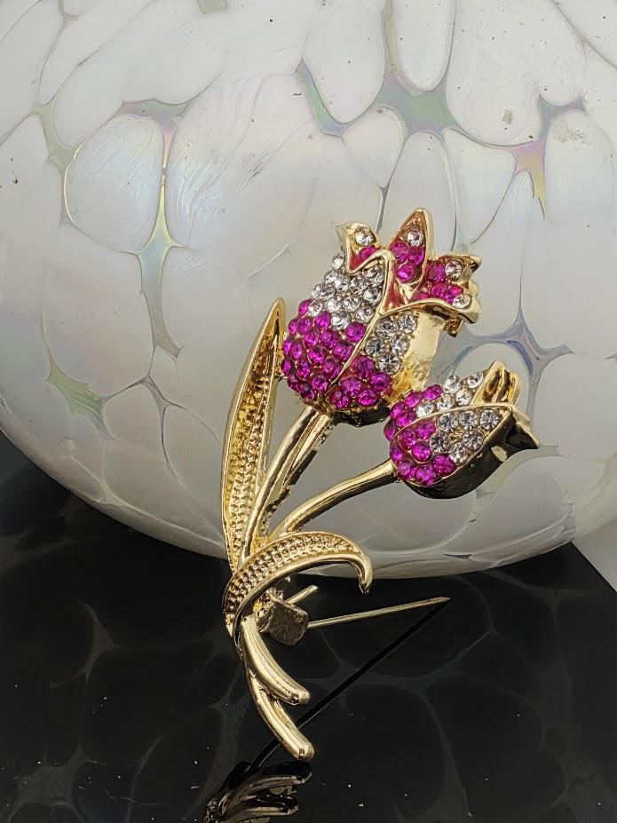 A decorative brooch in the shape of a flower, with pink and red gemstones, is displayed against a white and black textured background.