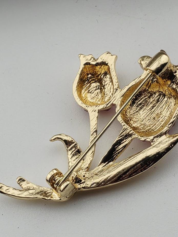 A close-up image of a gold-colored brooch with a floral design, featuring two open flower-shaped pendants.
