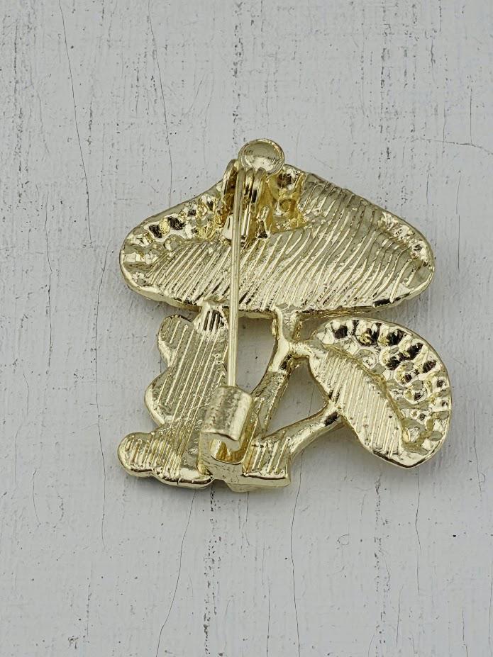 A gold-colored pin or brooch in the shape of a mushroom, with a leaf-like design on the stem.
