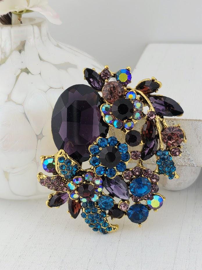 A decorative brooch with a variety of colorful gemstones and floral elements, including purple, blue, and brown stones, as well as some flowers.