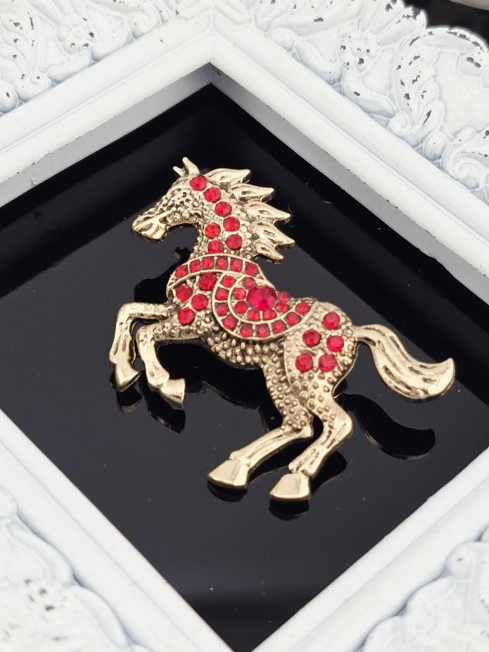A decorative gold horse brooch with red gemstones is displayed in a white ornate frame.
