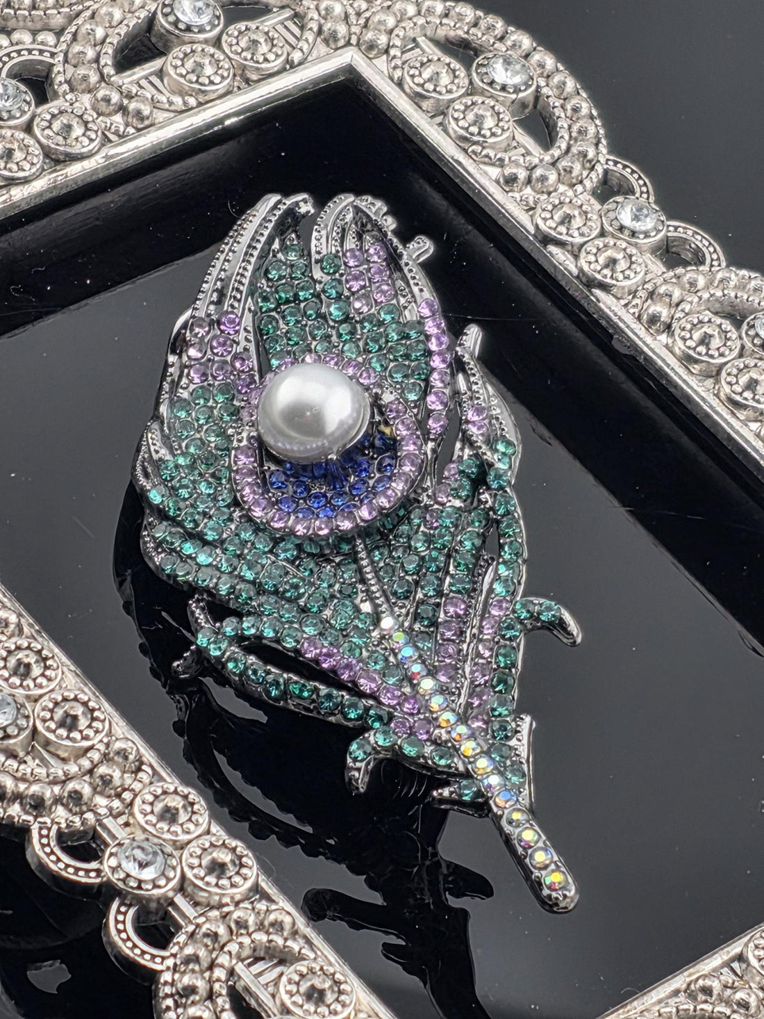 A close-up image of a decorative brooch with a feather-like design, adorned with colorful gemstones and a pearl at its center.