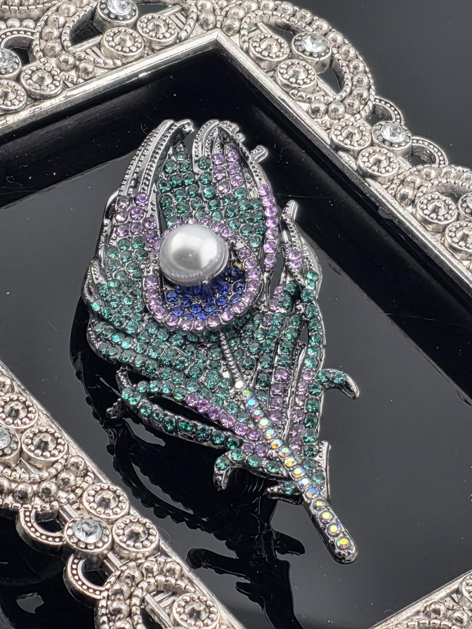A close-up image of a decorative brooch with a feather-like design, adorned with colorful gemstones and a pearl at its center.