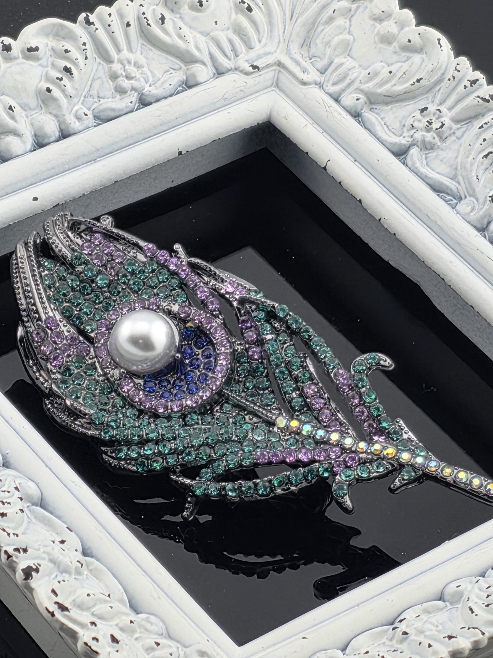 A decorative piece of jewelry featuring a large, colorful feather-like design with a pearl at its center, displayed within a white ornate frame.