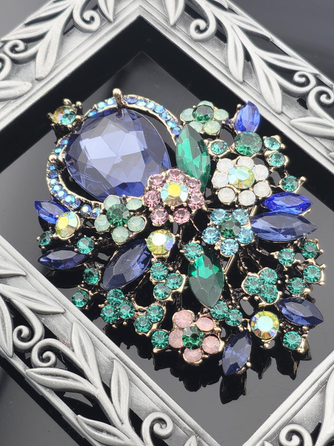 A decorative brooch with a variety of colorful gemstones, including blue, green, pink, and white stones, set within an ornate frame.