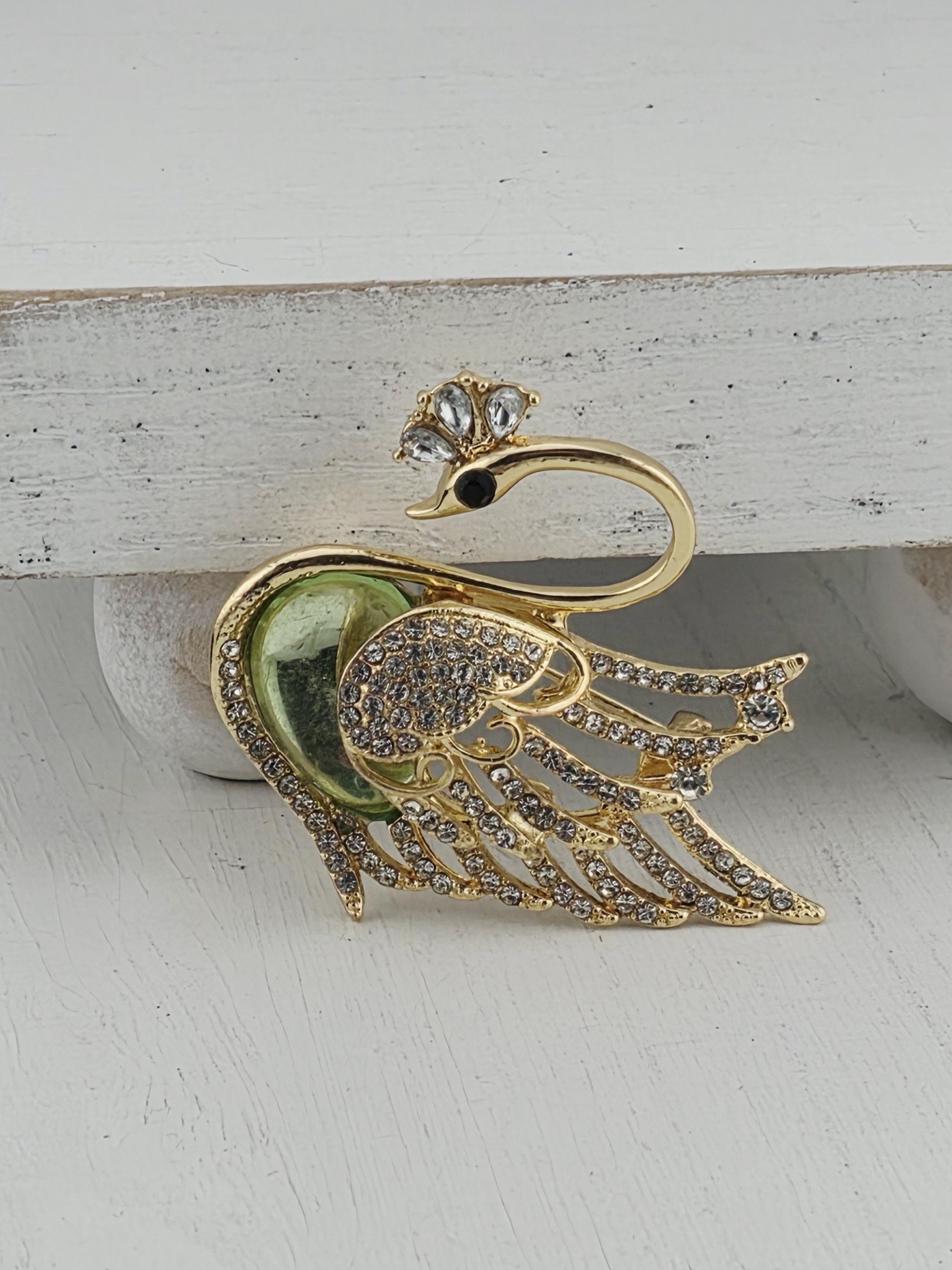 A decorative gold swan brooch with green and white accents, including a gemstone eye, is displayed against a white background.