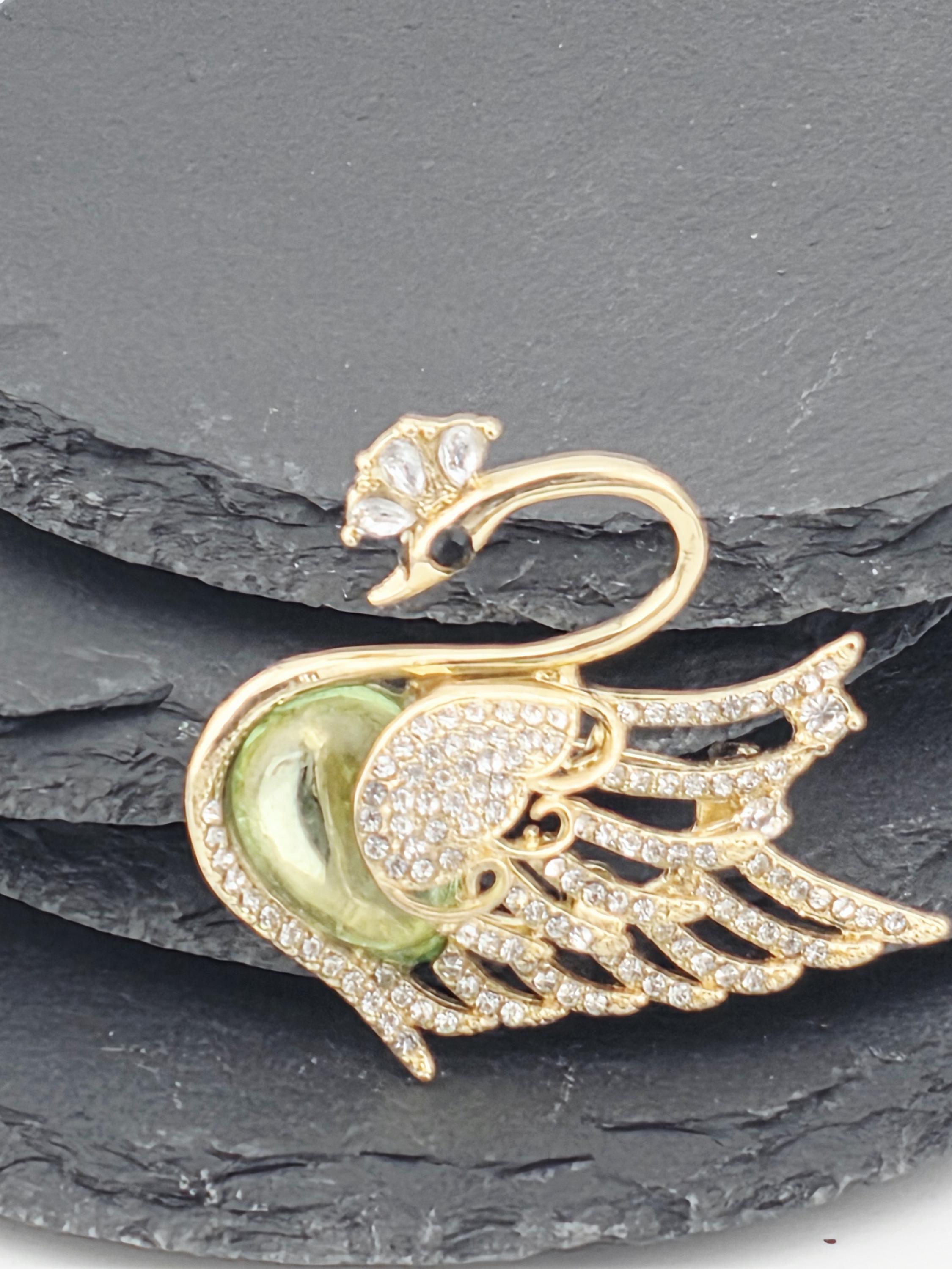 A gold-colored swan brooch with a green gemstone on its beak, set against a dark gray background.