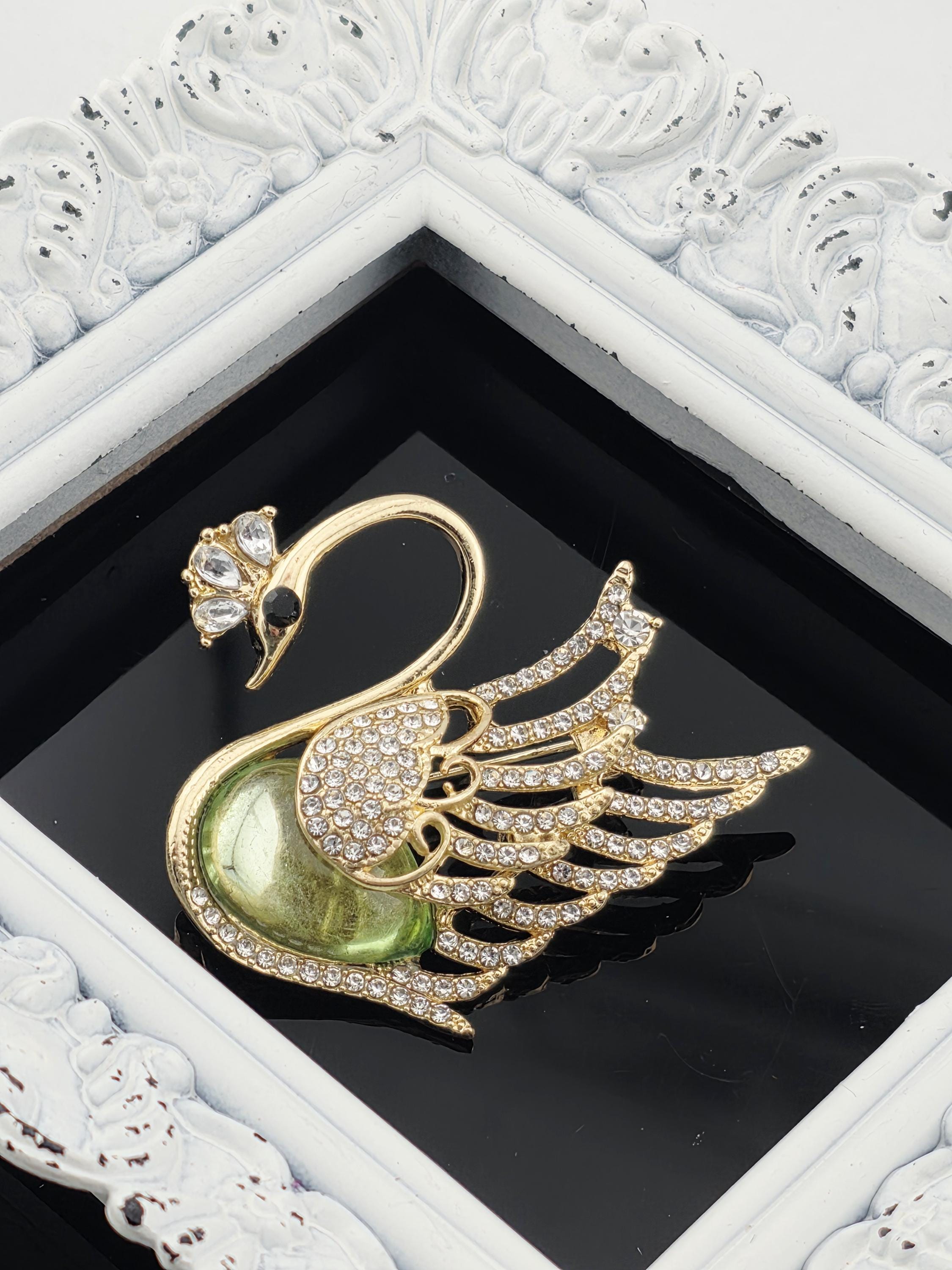 A decorative gold and green swan brooch with sparkling rhinestones is displayed in a white ornate frame.