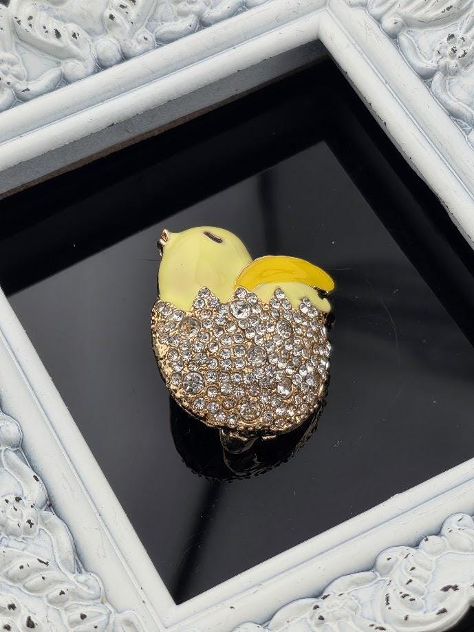 A decorative brooch in the shape of a yellow chick with a gold and crystal design, placed on a black background within a white ornate frame.