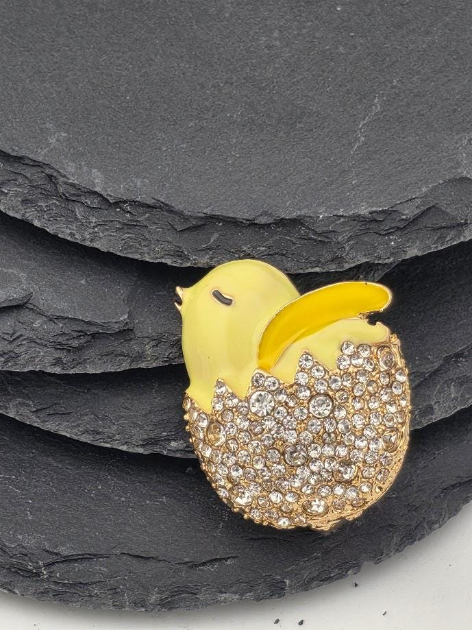 A decorative brooch in the shape of a yellow chick with a gold and crystal design, placed on a black background within a white ornate frame.