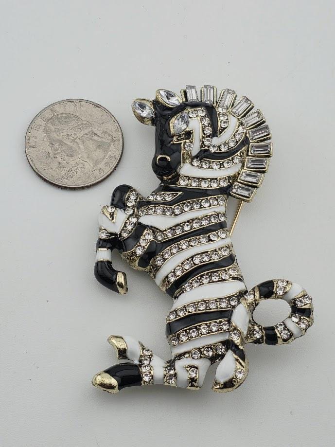 A decorative zebra brooch with a black and white striped pattern, adorned with rhinestones and a large rhinestone on its head. The zebra is positioned next to a coin, which appears to be a US quarter.