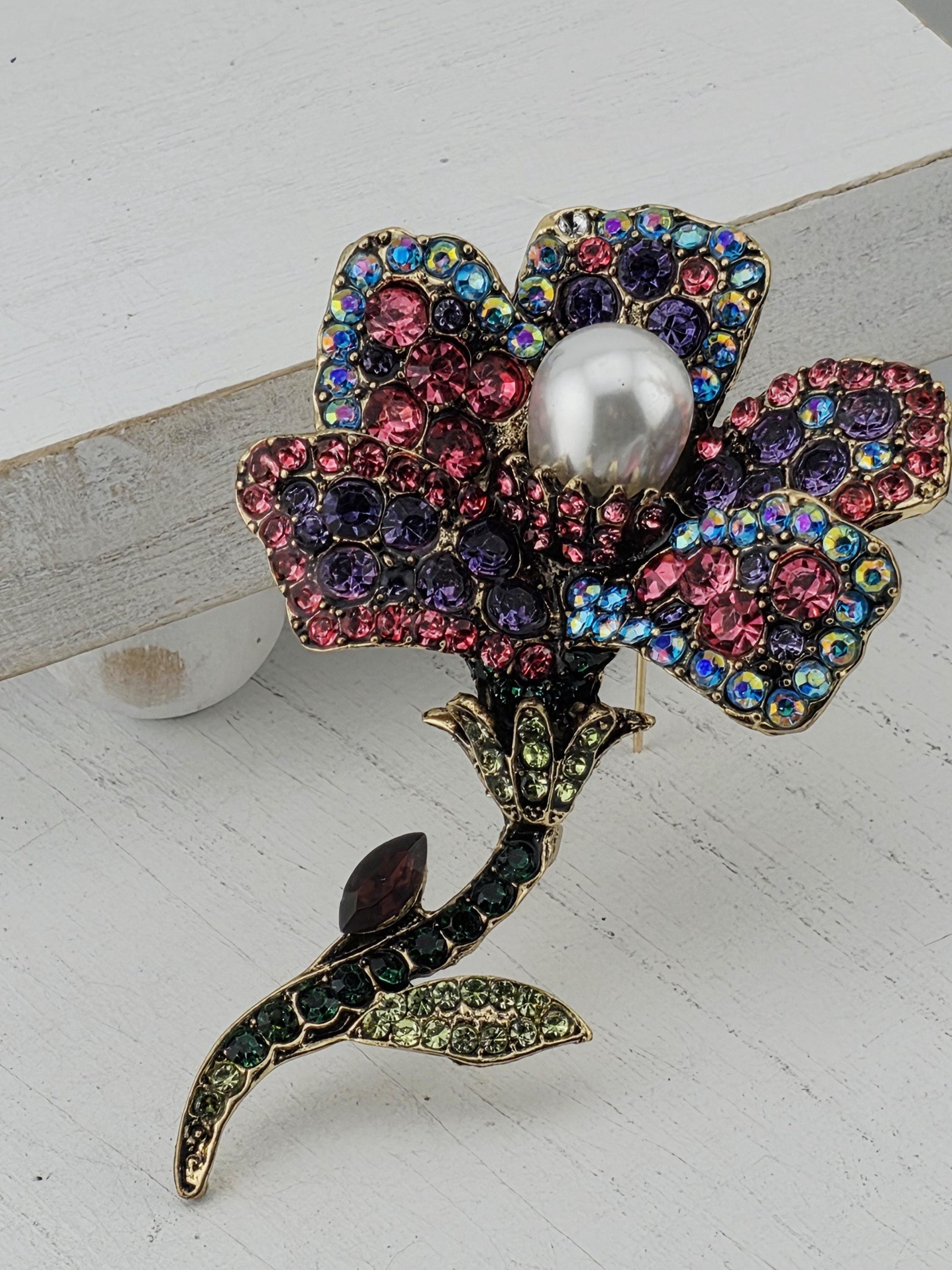 A decorative brooch with a flower-like design, featuring a variety of colorful gemstones and a large pearl at the center.
