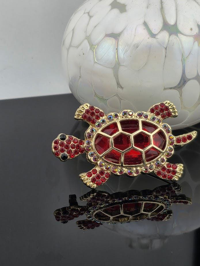 A decorative turtle brooch with red gemstones and gold accents, placed on a reflective surface.