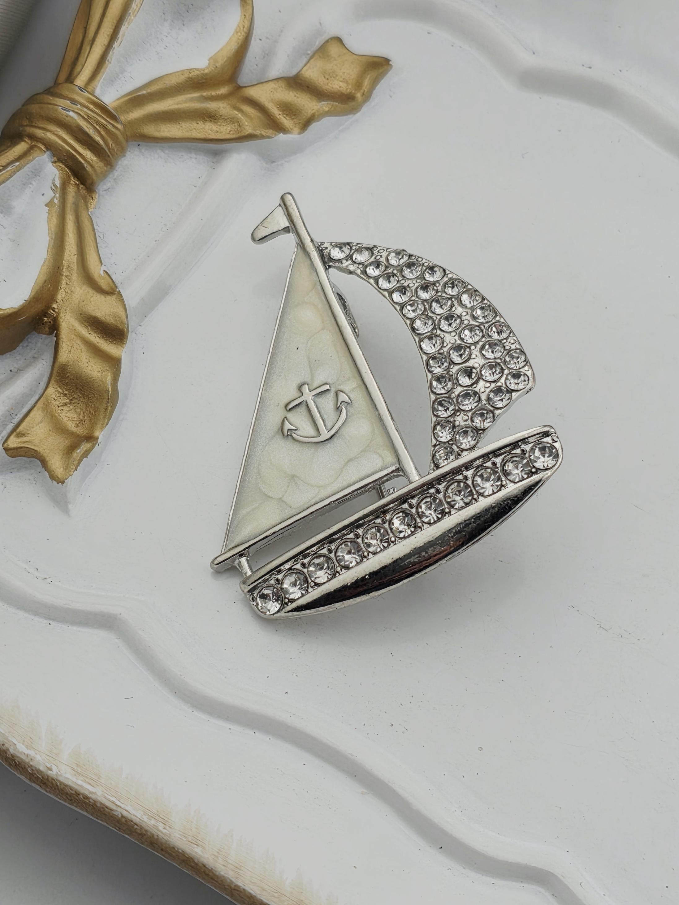 A silver and white brooch with a gold ribbon and a small anchor symbol is displayed on a white surface.